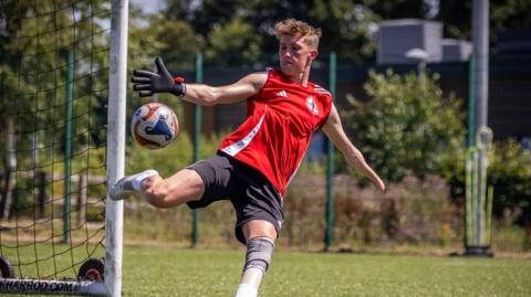 Thomas Atkinson, wearing a football kit with a red top, in action, about to kick a football away from his net.