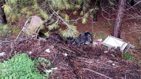 A haul of rubbish dumped at the bottom of a slope in a wooded area, There are wicker chairs with metal arms, a foldable white table and multiple black bin bags of household rubbish.