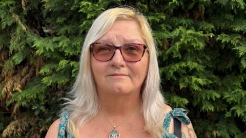 A woman with long blonde hair to her shoulders stands in front of some green trees. She wears sunglasses and a metal star-shaped necklace on a chain. The top of her dress straps, green with a paisley pattern, can just be seen