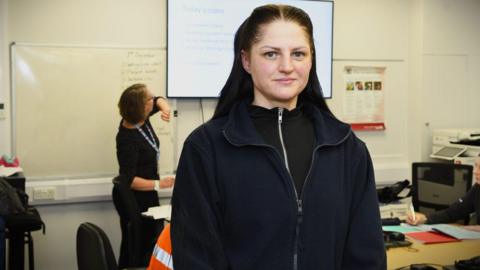 A woman with dark brown hair slicked back from her face but down at the back is looking into the camera. She is wearing a brown zipped fleece with a black zipped top underneath. Behind her is a classroom with someone sat down at a table and a woman with a lanyard looking at a screen.