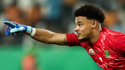 Ronwen Williams, wearing a red South Africa goalkeeping jersey and a blue glove on his right hand, leans forwards into shot from the right-hand side of the image and points with his index finger and arm extended as he looks ahead