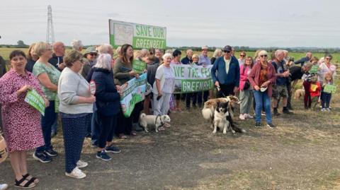 A group of protesters holding signs reading "Save our green belt". They stand next to the fields which are potentially affected. A number of people have dogs on leads.