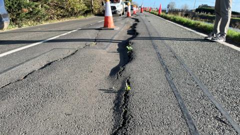 Part of the road with large, wavy cracks in the tarmac and raised sections. Orange and white cones stretch along the middle of the road as cars pass.