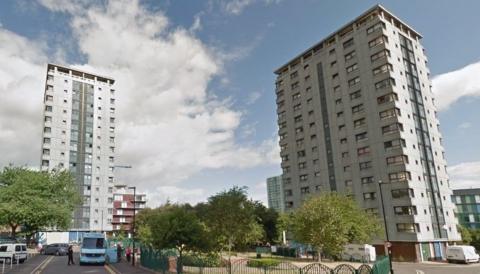 Grey blocks of flats under a blue sky with fluffy clouds. At the foot of the flats are several trees and a small fenced-off formal garden.