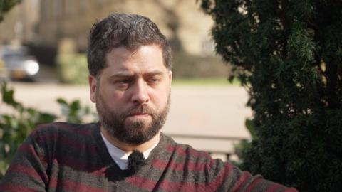 Kieron is a man in his 30s with brown hair and a brown beard He is photographed wearing a grey and red striped jumper over a white t-shirt. He is sitting on a park bench in front of a green bush.