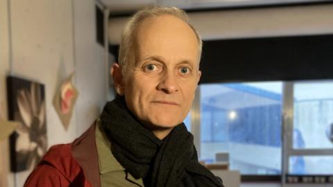 Dr Mark Wilsher, a man with short grey hair. He is looking into the camera and is smiling. He is wearing a khaki green shirt with a red coat and black scarf. He is stood inside his art studio. 