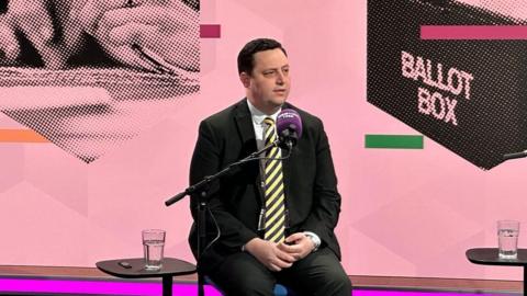 Ben Houchen is sitting in front of a ˿ Radio Tees microphone. He is wearing a black suit and yellow-striped tie. The graphic behind him show a ballot box.