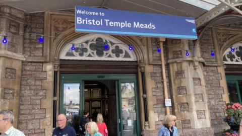 The entrance to a stone building. A blue sign reading Bristol Temple Meads Station hangs above. Members of the public can be seen in the foreground. There are pots of p9ink geraniums on the right.  