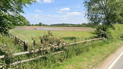 A body of water in the middle of a muddy field, surrounded by trees and grass by the side of a road.