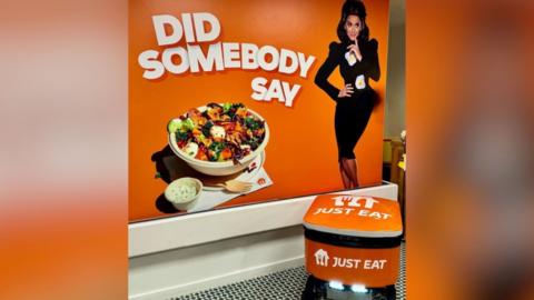 A photo of a Just Eat poster and a food delivery robot. The poster has an orange background with text that says "Did Somebody Say". Also on the poster there is a woman in a black dress with fried eggs on her dress. There is also a bowl for food with a Wooden fork and a pot of white sauce. The Just Eat robot is orange, in the shape of a box, with wheels underneath