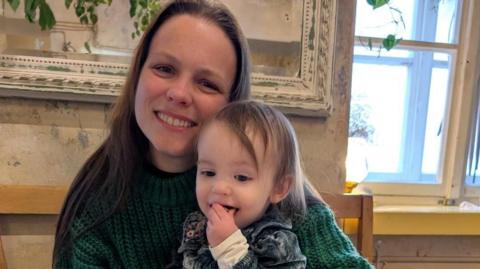 A mum and daughter in a room with limewashed walls and a mirror behind. They are sitting on a bench, the child on the mum's knee. The mum has long brown hair and is wearing a green chunky knit jumper. The child, who is a toddler, has her hand in her mouth as she eats something and has short brown hair and is wearing a denim dress with floral collar. Both are smiling, the mum looking at the camera while the child looks down.