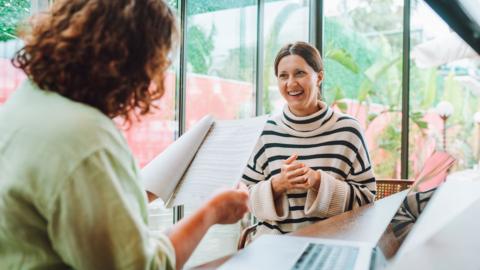 A manager reads the resume of an interview candidate, who is sitting across from her, wearing a black and white striped top