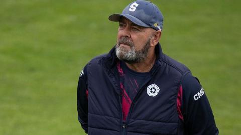 Darren Lehmann walks around the ground with his arms behind his back looking up at the stand.