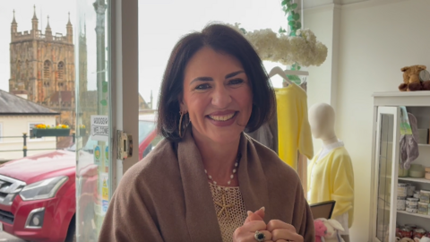 A woman with a darl brown bob is wearing hoop gold earrings a pearl necklace with a starfish pendant and has a brown shawl on. She appears to be standing in a clothes and gift shop