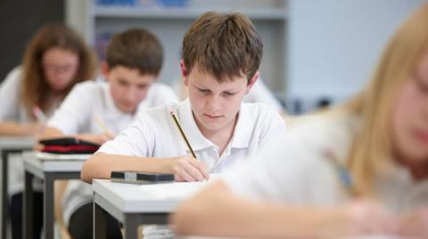 School children wearing white polo shirts sitting at desks in a row. The focus is on a boy who has brown hair and is writing with yellow and black striped pencil