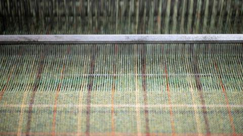 A green tweed being woven on a loom. There is pattern of brown, orange and yellow through the fabric