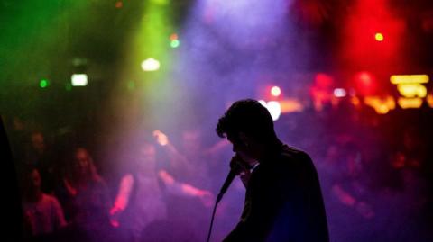 A member of the French beatbox collective EOG performs during the New Urban Era Beatbox Battles competition, at the ATIK nightclub in Tamworth in 2023. The man can be seen in a dark silhouette with colourful lights behind him as well as members of the crowd. 