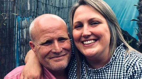 Laura Barlow has her arm around her husband Mike. She has shoulder-length hair, is smiling broadly and wears a black-and-white checked shirt. He has short hair and wears a pink shirt.