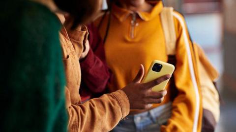 A close up of three people looking at a phone in a yellow phone case. The students are wearing brightly coloured clothes. 