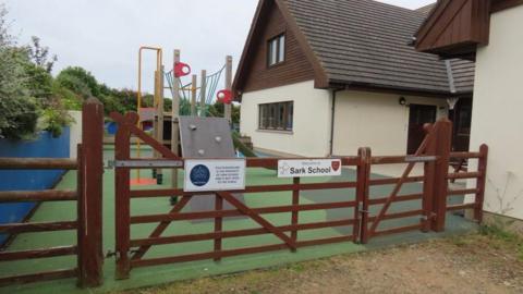 A wooden gable gate with a Welcome to Sark School sign. Behind a play area with climbing ropes, a green AstroTurf floor and a modern single-story school building with cream walls and a tiled roof.