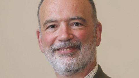 John Denerley, has short, grey/black hair and a mainly grey beard. He is wearing a white checked shirt