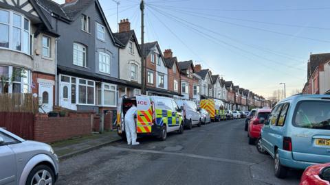 A row of houses and parked cars in a residential road with two police vans outside. A person wearing a forensic suit can be seen from the back at the back of one of the police vehicles.