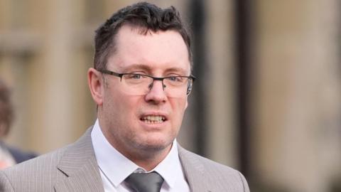 Image shows man in a light-brown suit with a silver tie and black-rimmed glasses. He has black hair and grey stuble.