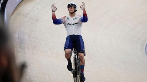 Matt Richardson riding a bike on a wooden track. He is wearing a white, blue and red cycling outfit and holding up his hands. 