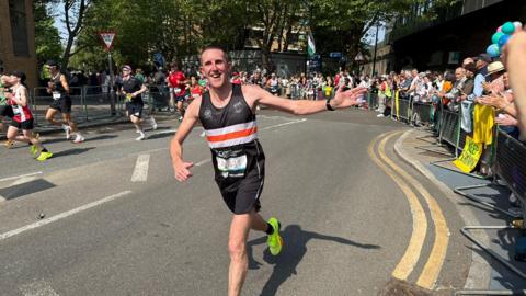 A man in a black, white and orange running vest with race number visible runs along a London street on a sunny day, reaching out towards spectators who line metal barriers on the right. Other runners are visible behind him and trees are in full leaf overhead. 