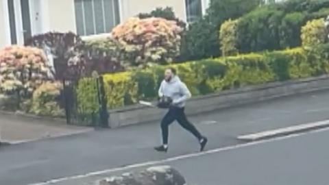 Man running down street with chainsaw. He is wearing a grey hoody and black trousers.