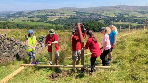 Photograph of volunteers installing leaky dams on Holcombe Moor. The image shows wooden posts being hammered into a gulley on the hillside.