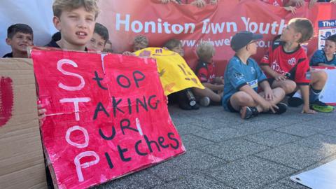 Children sat protesting. They are holding banners and painted signs. One reading "Stop taking our pitches".
