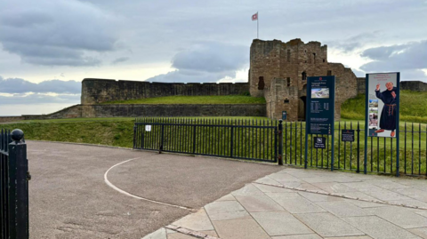 The long, low, black metal gates leading to Tynemouth Priory and Castle are open. The ruins of the stone building, which includes a tower and an English Heritage flag, are in the background across an area of grass. Two tall signs are by the metal fence, one showing opening times and the other containing a cartoonish painting of a jolly monk.