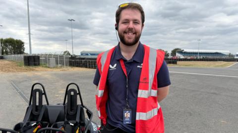 A man wearing a red high vis jacket smiling at the camera standing next to a black go kart with two seats. He is located on a karting track, with a wall of tires behind him.