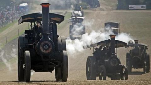 Six steam engines travelling up and down a hill across a field towards the camera with crowds seen to the left