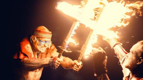 Two men in battle with Celtic inspired face paint and costume and fire torches. 
