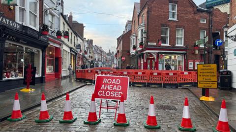 The roadworks in Bailgate, Lincoln. There is a line of red and white roadwork cones across the cobbled street. Above them is a red and white sign reading ROAD AHEAD CLOSED
