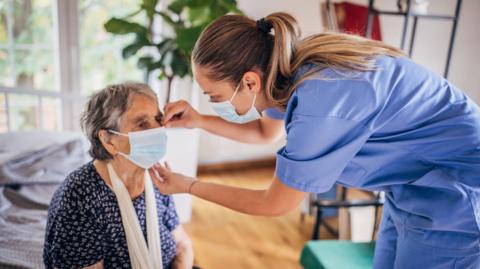 A nurse wearing scrubs and with a face mask on checks an elderly woman, also wearing a face mask, in a nursing home