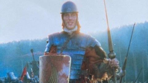 Scot Newing dressed in armour, carrying a shield, in front of the woods and other extras 