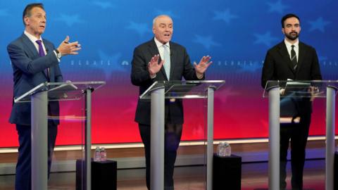 Andrew Cuomo, Curtis Sliwa and Zohran Mamdani stand at glass podiums on a stage with red and blue starred background and small stools with water next to them
