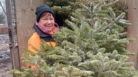 A woman in a high-visibility orange jacket and a black beanie hat is loading a tree onto a the back of a lorry. She is looking at the camera, smiling.