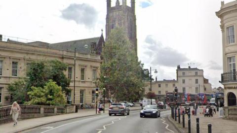 A street with beige-coloured buildings and a church on the left hand side with black bollards lining the left side of the road, cars on the road and people walking on the pavement
