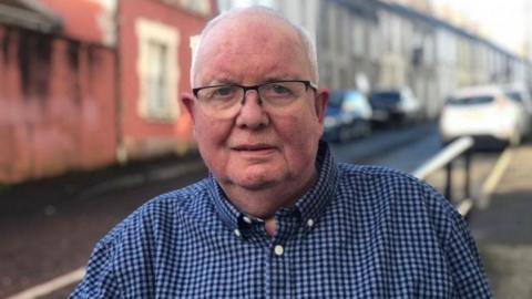 Patrick Gill stands on a street. There are cars and terraced houses behind him. He has grey hair and wears glasses and a blue checked shirt. 