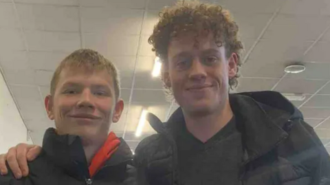 Two young men smile at the camera - Charlie on the left has short blonde hair and is wearing a black coat and a red hoodie. Oli is taller and has his hand on Charlie's shoulder. Charlie has curly hair and is wearing a black jumper and black coat. 