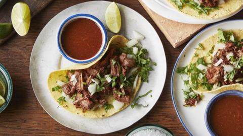 Soft taco filled with beef, chopped onion and coriander on a plate with a lime wedge and salsa sauce