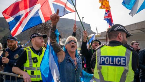 A blonde-haired woman wearing blue eye make-up points in the air as she chants outside the Scottish Parliament building in Edinburgh. She has a police officer on either side of her.