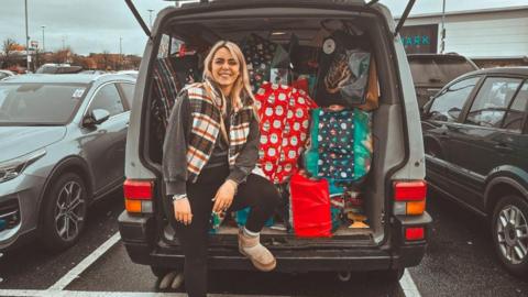 Katie Tradie sits on the edge of her open boot car, full of bags of supplies, with one foot on the back bumper. She wears low rise uggs and a checked gilet. She has blonde hair and smiles at the camera. 