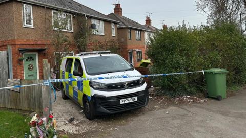 A police van is parked on the drive of a house. Police tape stretches across the drive from a green bin to a fence panel. Some flowers have been laid to the left of the drive, by the fence panel. The front door of the house is green with red brick around the lower level of the house it and brown render above.