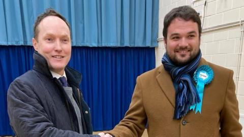 Scott Hussey is wearing a dark blue coat over a white shirt, blue tie with white spots and a grey jumped. To his right, Robin Hunter Clarke is wearing a brown overcoat, blue striped jumped and blue Reform UK rosette.