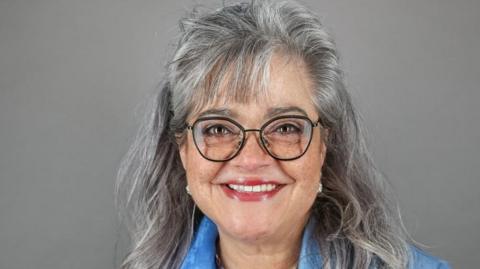 A woman with long grey hair and glasses in front of a plain grey backdrop smiles at the camera.